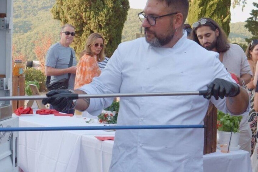 A person in a white chef's coat holding a long metal rod, likely for cooking or serving. 1