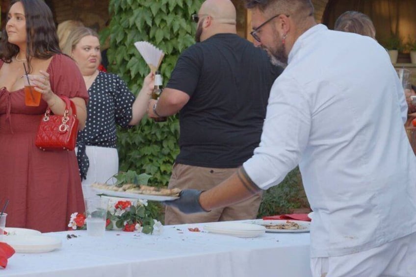 A group of people dining outdoors with a table set for a meal, surrounded by greenery. 4