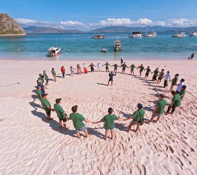 A group of people forming a circle on a sandy beach with boats in the water and mountains in the background. 2