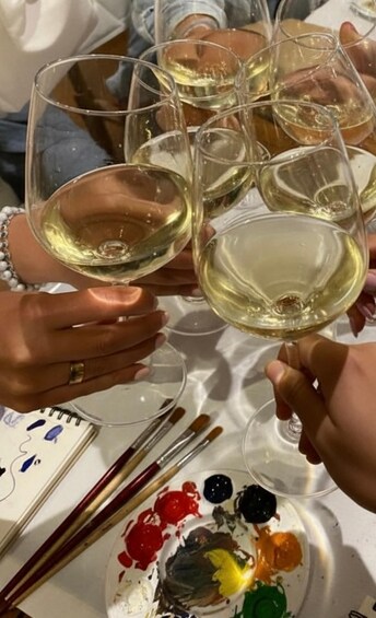 People toasting with white wine glasses, a paint palette with various colors, and paintbrushes on a table. 5