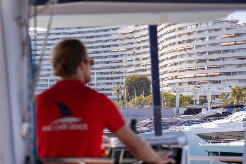 Une personne portant un t-shirt rouge orné d’un logo, aux commandes d’un bateau dans une marina où se trouvent plusieurs yachts, avec un grand bâtiment en arrière-plan. 1
