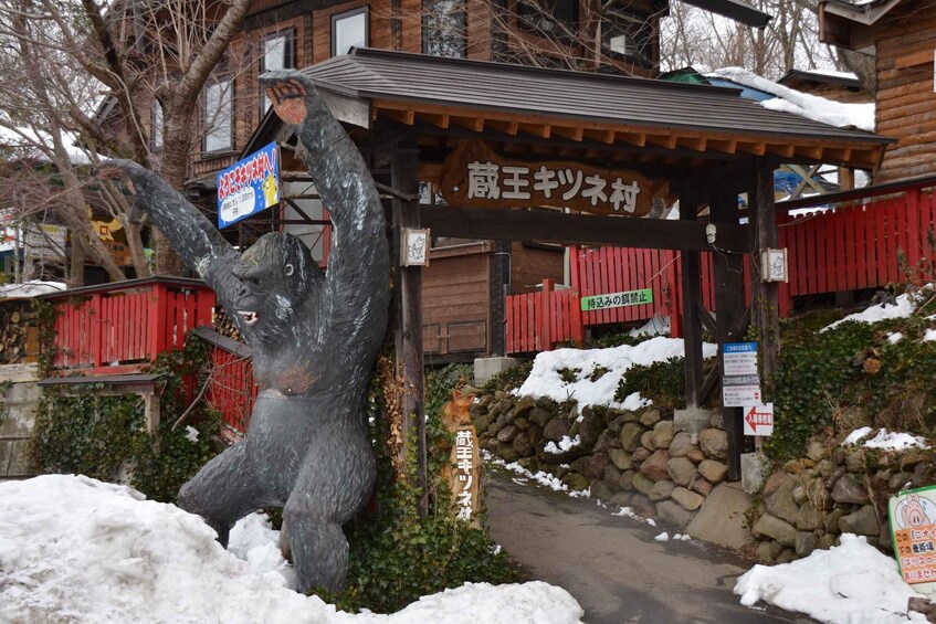 A snow-covered statue of a bear, a wooden sign with Japanese text, and a red fence. 4