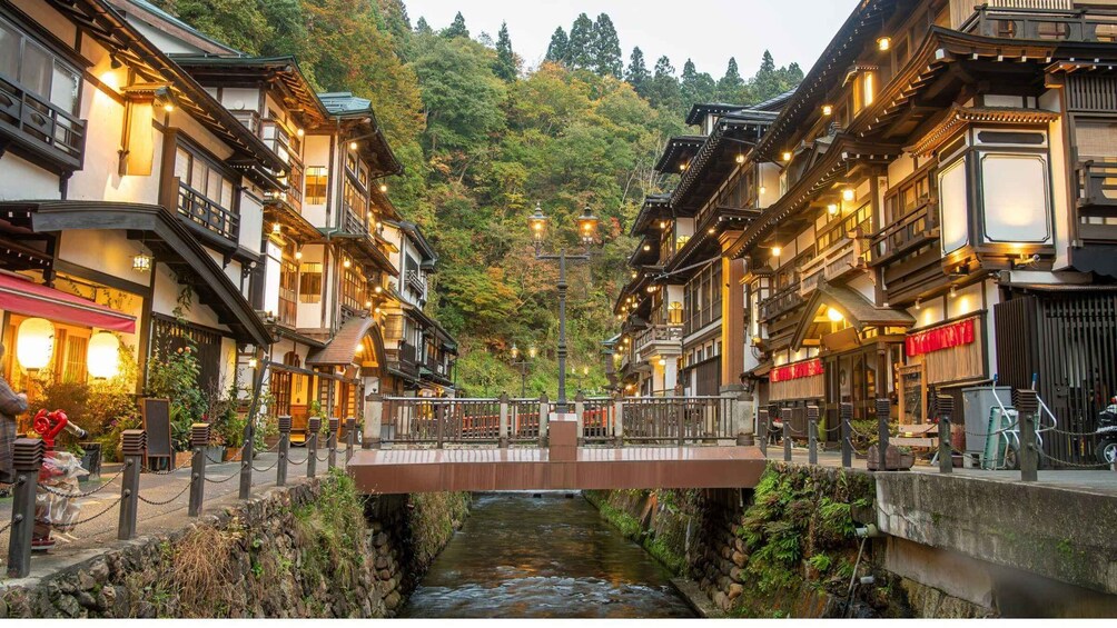 A traditional wooden bridge over a narrow stream in a mountainous area with buildings on either side. 1