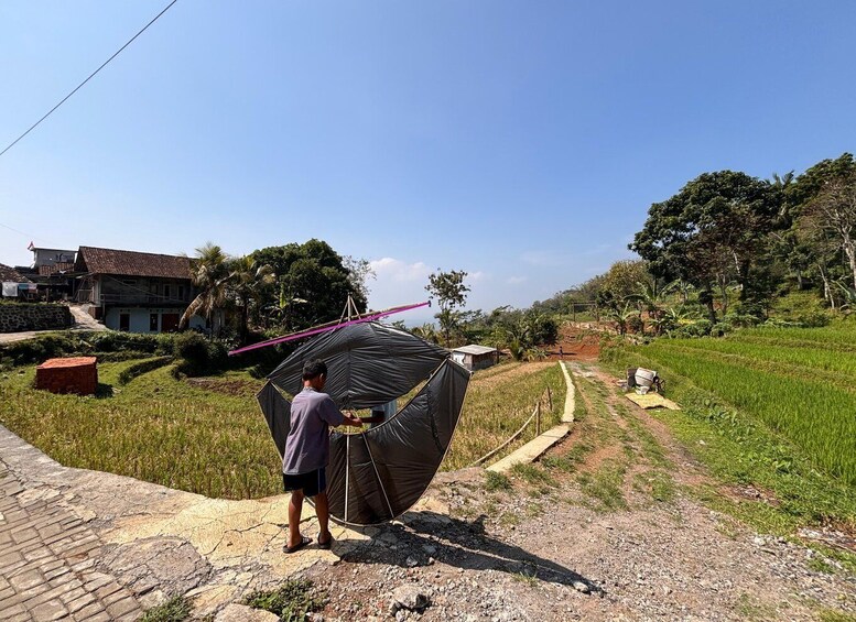 A man setting up a large black tent on a rural road with a house and fields in the background. 4