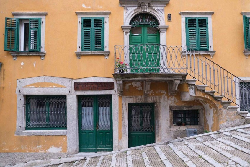 A yellow building with green shutters, a balcony with a wrought iron railing, and a cobblestone street. 1