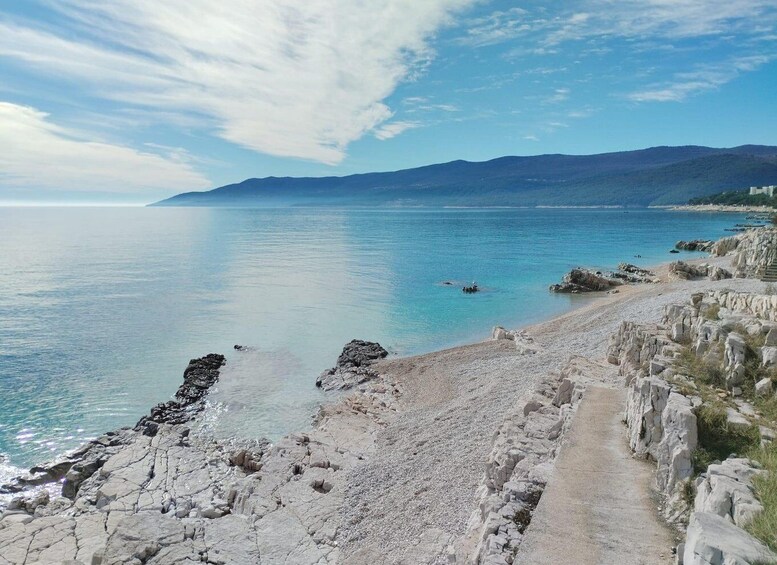A coastal landscape with a rocky shoreline, clear blue sea, and distant mountains under a partly cloudy sky. 3