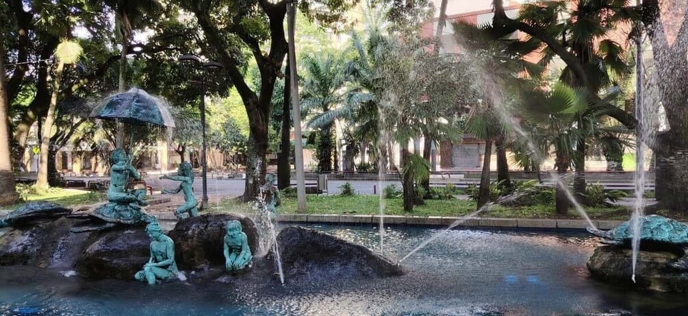 A fountain with statues and water jets, surrounded by trees and a paved walkway. 3