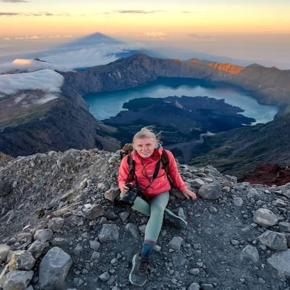 A person sitting on a rocky mountain peak with a lake and mountains in the background. 1
