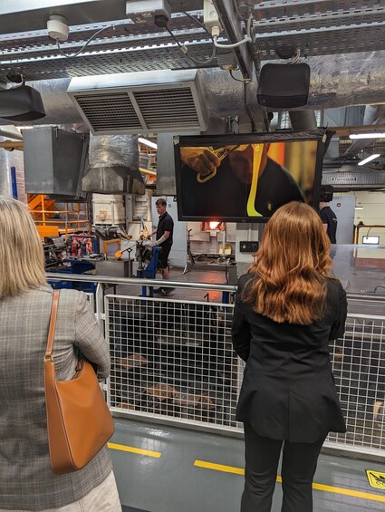An industrial setting with machinery, a large screen displaying a video, and two individuals observing. 5