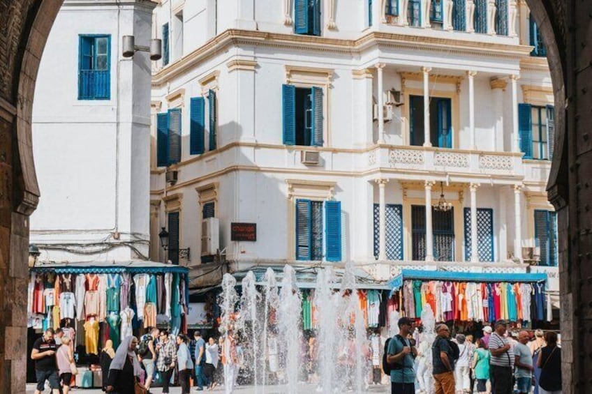Een levendige markt met een fontein in het midden, omringd door witte gebouwen met blauwe luiken. 3