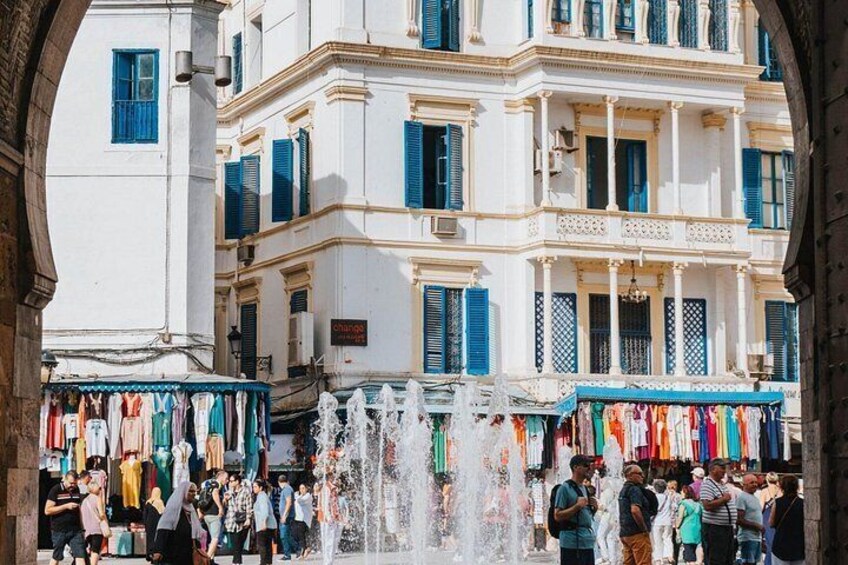 Un mercato vivace con una fontana, edifici bianchi con persiane blu e persone che fanno acquisti. 2