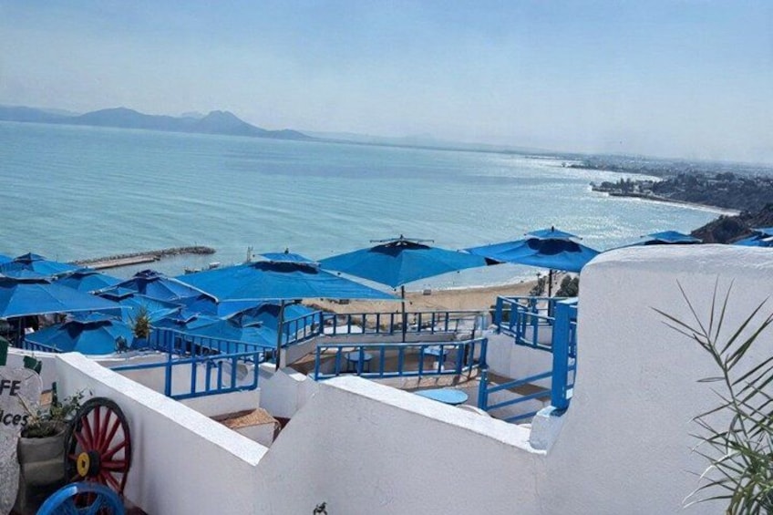 Een terras aan zee met blauwe parasols, een wit gebouw en uitzicht op de zee en verre bergen. 1