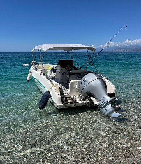 A white motorboat with a canopy, anchored in clear turquoise water with a mountainous coastline in the background. 3