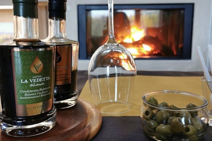 Two bottles of La Vedetta Balsamic Condimento, a glass bowl of olives, and a wine glass on a wooden board. 2