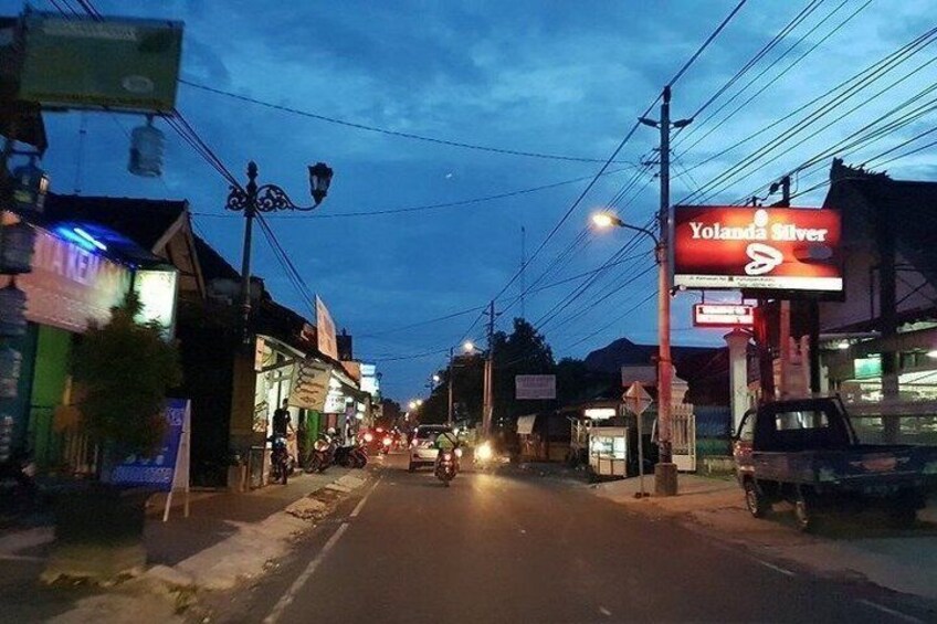 A street at dusk with illuminated signs, including 'Yolanda Silver,' and a motorcycle in the distance. 2