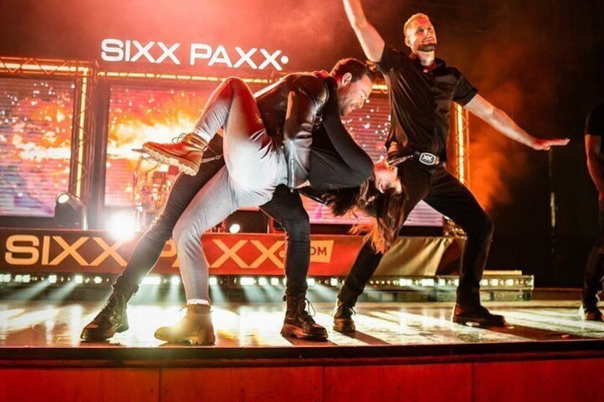 Une scène où des artistes adoptent des poses dynamiques, un grand écran affichant le texte « SIXX PAXX », et un décor rouge et jaune. 3