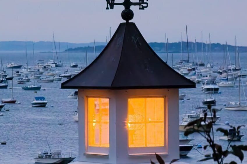 A lighthouse with a lit window overlooking a marina filled with sailboats. 5