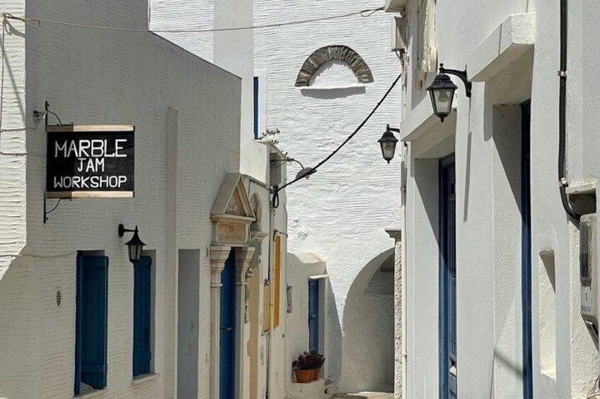 Une rue étroite bordée de bâtiments blancs, de volets bleus et d’une enseigne indiquant « ATELIER DE FABRICATION DE CONFITURES DE MARBRE ». 2