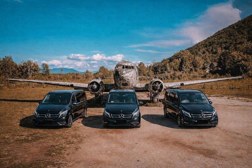 Three black Mercedes-Benz SUVs parked in front of a large aircraft on a dirt ground with a mountainous backdrop. 3