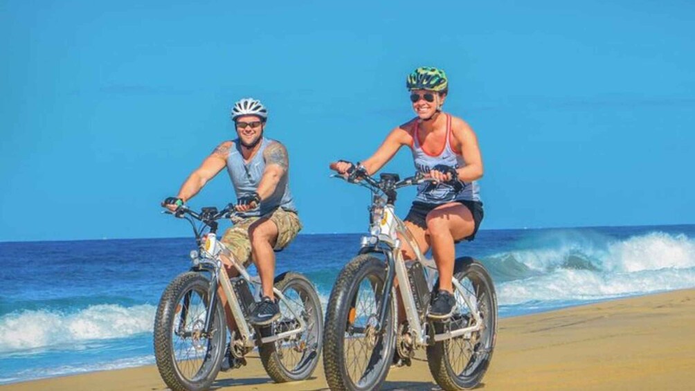 Two people riding bicycles on a beach with the ocean in the background. 5