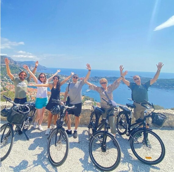 A group of people on bicycles with their arms raised, overlooking a coastal landscape with buildings and boats. 1