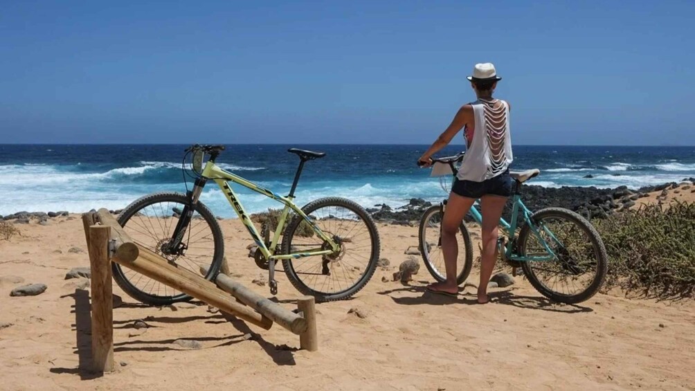 A person standing on a sandy beach with two bicycles, one leaning against a wooden post, and the ocean in the background. 2