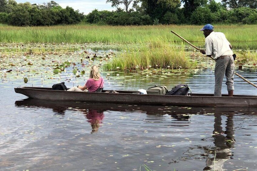 A person standing in a boat on a body of water with another person seated inside. 1