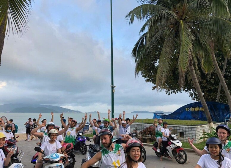 A group of people on motorcycles, some wearing helmets, raising their hands in the air, with a body of water and palm trees in the background. 3