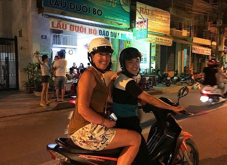 Two people on a motorcycle, a street with shops, and a sign with the number 93. 2