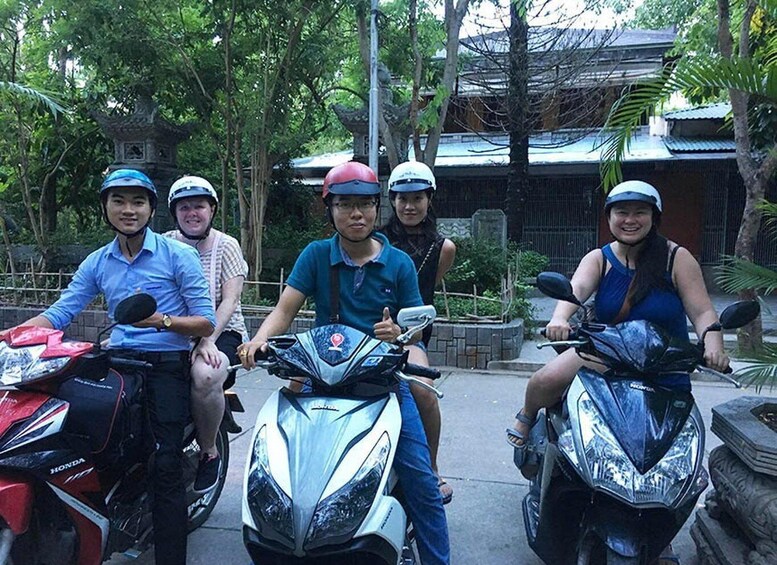 Four people on scooters in a tropical setting with traditional architecture. 5