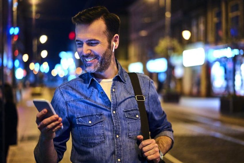 Una persona parada en una acera de noche, sosteniendo un teléfono inteligente y vistiendo una camisa vaquera con una mochila. 2
