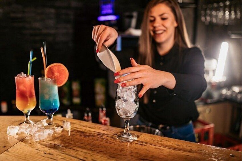 Un barista versa un drink in un bicchiere con cubetti di ghiaccio, con due cocktail colorati sul bancone. 1