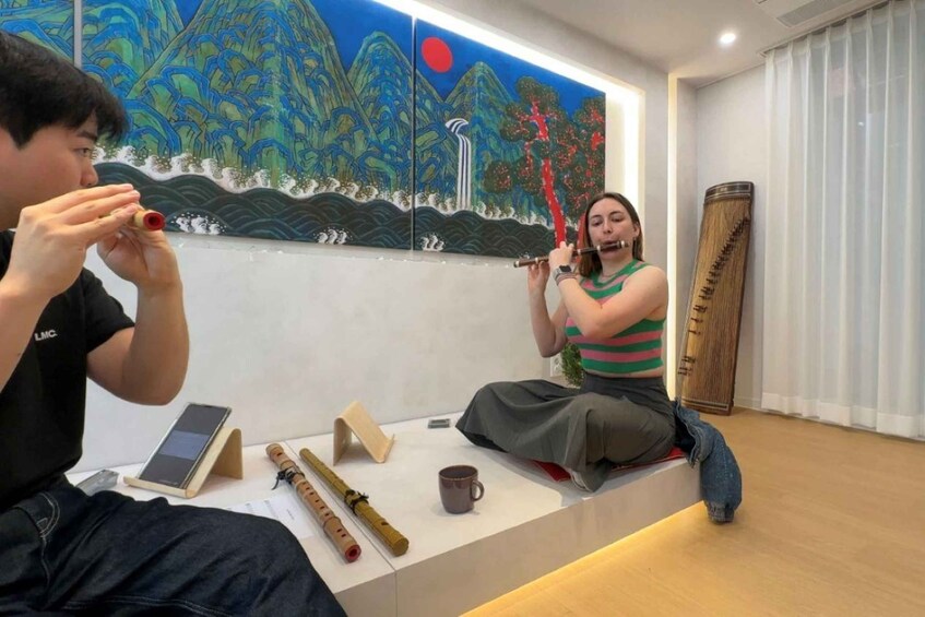 Two people playing traditional flutes in a room with a mural of a mountain landscape. 2