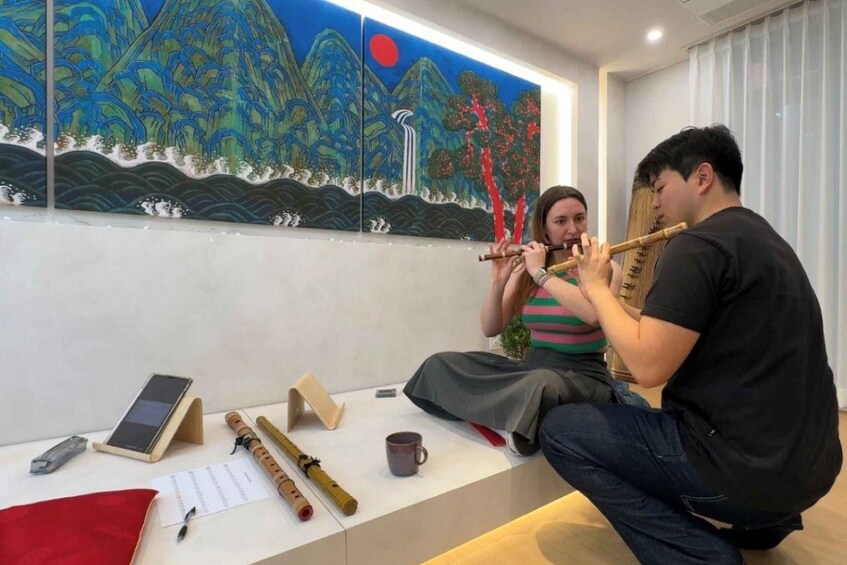Two people playing traditional flutes in a room with a large painting of a mountain landscape. 3