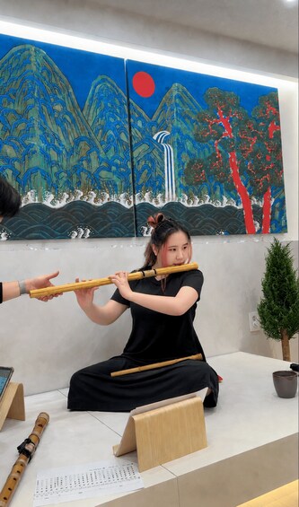 A person playing a traditional flute, seated in front of a wall with a painting of mountains and a red circle. 5