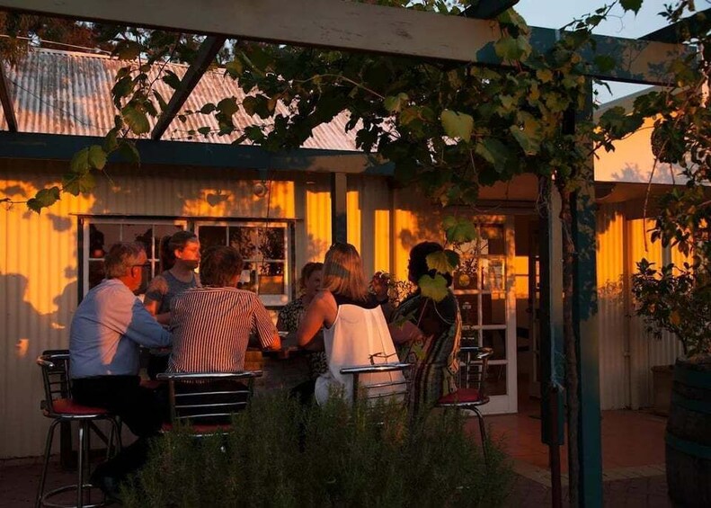 A group of people seated at outdoor tables under a pergola, with a building in the background. 4