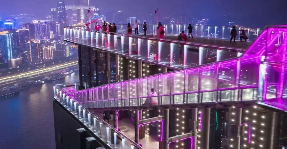 A brightly lit pedestrian bridge with glass railings overlooking a cityscape at night. 3