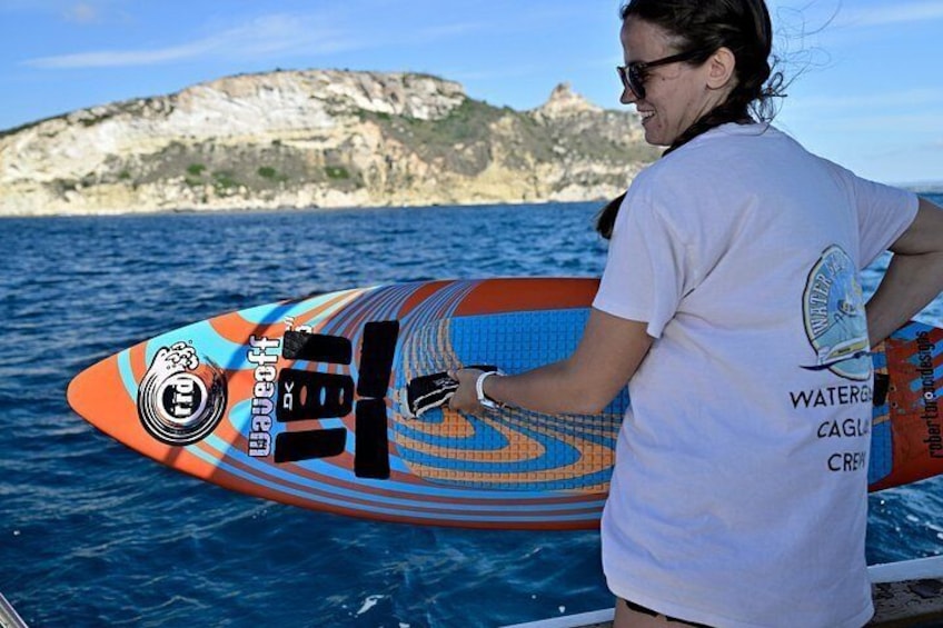 A person holding a colorful surfboard with a logo and text, standing on a boat with a rocky coastline in the background. 3