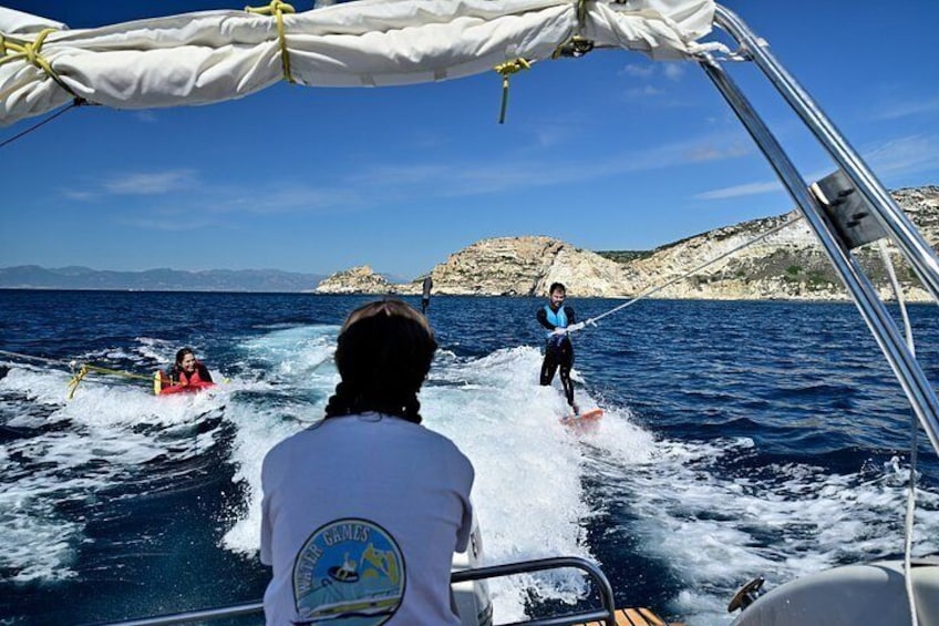 A person wakeboarding on a boat, another wakeboarding, and rocky hills in the background. 2