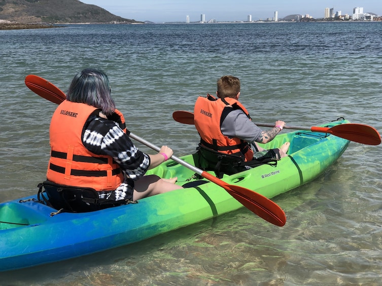 Two people kayaking on a calm body of water with a city skyline in the background. 2