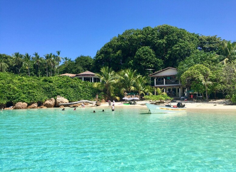 A tropical beach with clear turquoise water, a boat, and a resort with palm trees and a mountain backdrop. 1