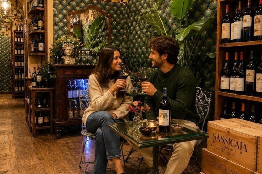 Due persone sedute a un tavolo in una cantina vinicola, con scaffali pieni di bottiglie e una scatola con l'etichetta 'SASSICAIA'. 2