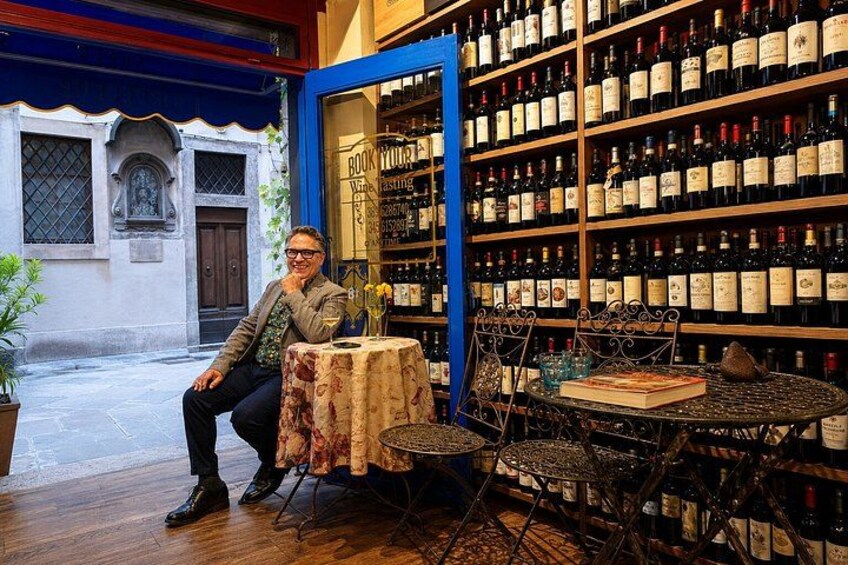 A man seated at a table with a wine bottle and glasses, in front of a wine cellar with numerous bottles. 4
