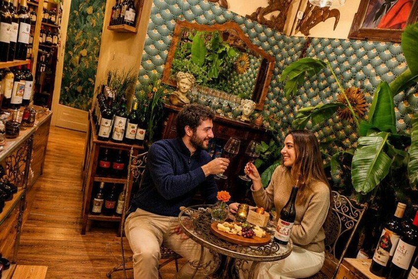 A couple dining in a room with a patterned wall, wooden shelves with bottles, and a large plant. 3