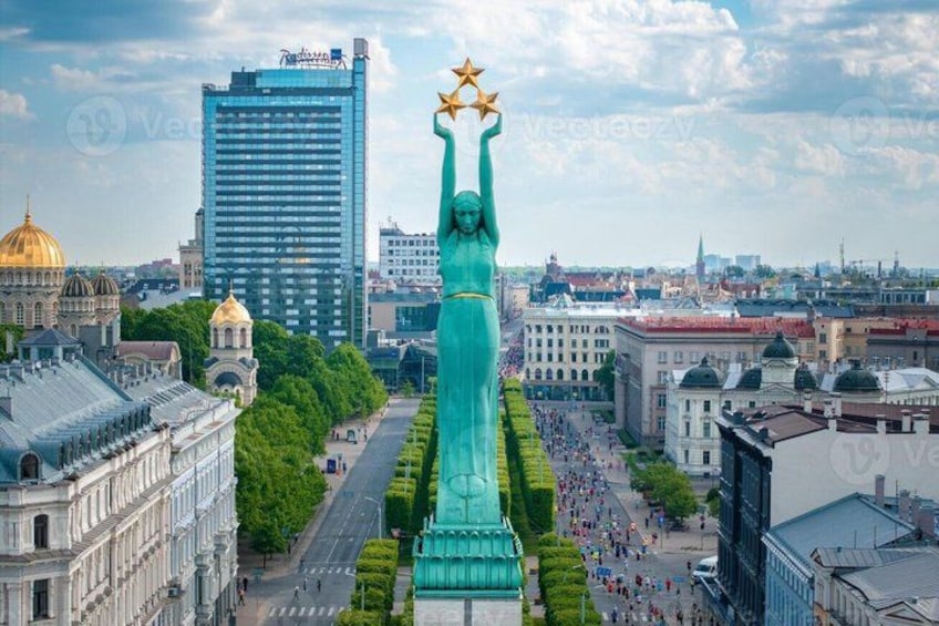 A large statue with a star-topped head, surrounded by urban buildings and a tree-lined street. 1