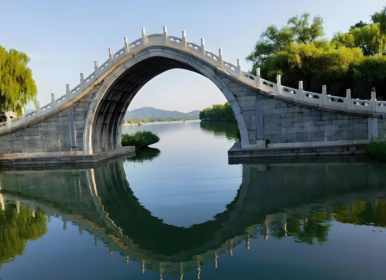 A stone bridge over a calm river with a reflection, surrounded by trees and distant mountains. 5