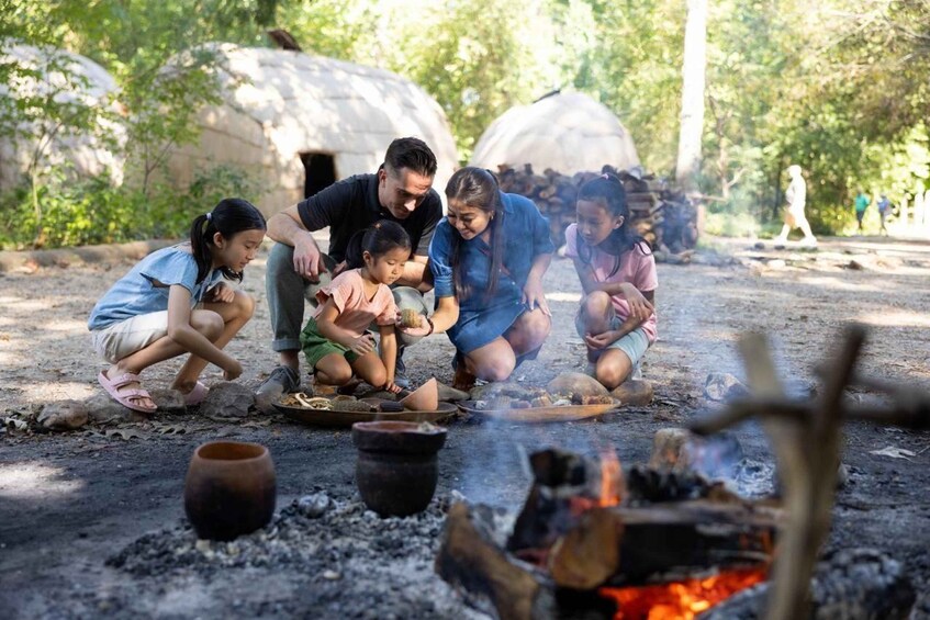 A group of people around a campfire with pottery and food, in a natural setting with trees and a thatched-roof structure in the background. 4