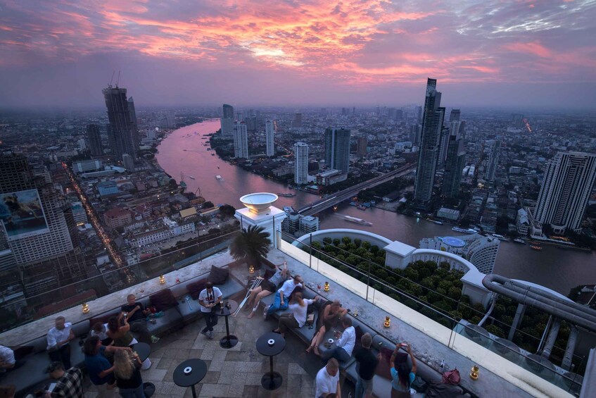 A cityscape at sunset with a river, high-rise buildings, and a rooftop bar with people. 3