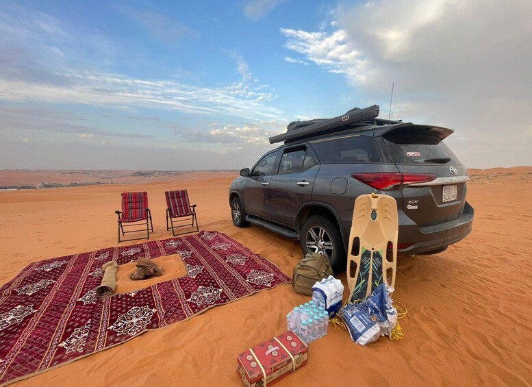 A desert landscape with a parked SUV, a picnic setup with a blanket, two chairs, and camping gear. 2
