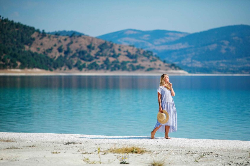 A woman in a striped dress walking on a beach with a calm lake and hills in the background. 1